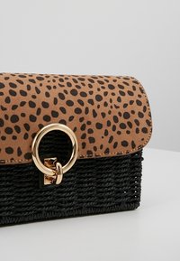 Sac tan avec un motif à pois noirs en haut et une base tissée noire. Dispose d'un fermoir circulaire en or et d'un design structuré.