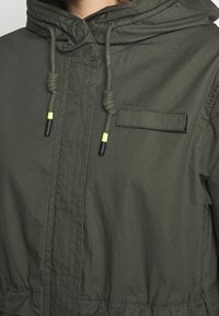 Chaqueta con capucha de color verde oliva que cuenta con un bolsillo frontal, cordón ajustable con detalles amarillos y una pretina ceñida. Textura lisa y mate.