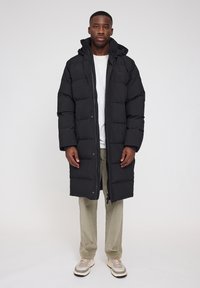 Schwarze Daunenjacke mit Reißverschluss- und Druckknopfverschluss, ausgestattet mit einer Kapuze und gestepptem Design. Getragen über einem weißen Hemd, kombiniert mit hellen Hosen und Turnschuhen.