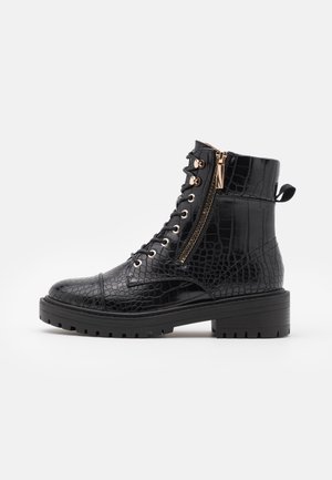 Zwarte enkelboots van glanzend krokodillen-geprofileerd materiaal. Voorzien van gouden hardware, veterdesign aan de voorkant, zijrits en een chunky rubberen zool.