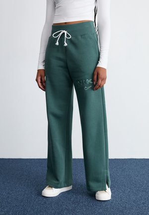 Mujer con pantalones deportivos verdes de Nike con cordón blanco, camiseta blanca de manga larga recortada y zapatillas blancas sobre alfombra azul.