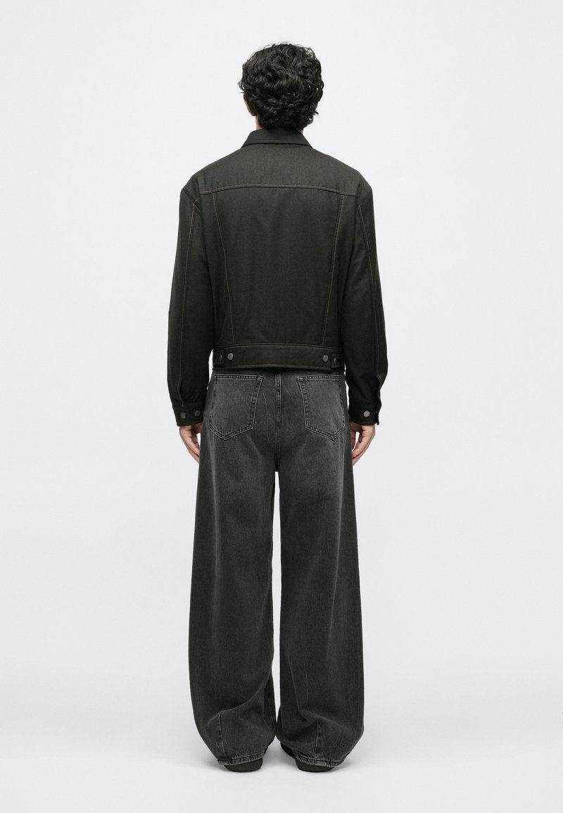 Sort denimjakke med synlige sømme og knapper, parret med løse, mørk grå denimbukser. Outfittet har en afslappet pasform og minimal pynt.