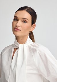 Blouse blanche en tissu léger, avec un col haut et un détail de cou noué, des manches bouffantes et une silhouette structurée.