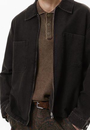 Une personne porte une veste noire non zippée par-dessus un polo marron en tricot, un pantalon à motif paisley et une ceinture marron avec une boucle argentée.