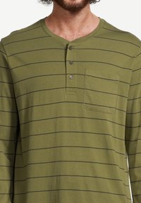 Mies, který má na sobě olivově zelenou dlouhý rukáv henley košili s tenkými černými vodorovnými pruhy a kapsou na přední straně hrudi, zapnutou u límce.