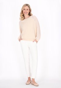 Felpa beige con maniche in pizzo, abbinata a pantaloni bianchi slim-fit e scarpe beige slip-on. Tessuto morbido, vestibilità rilassata e design leggero.