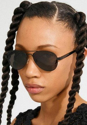 Gafas de aviador negras con lentes tintadas y monturas metálicas. Cabello peinado en dos trenzas retorcidas. Piel iluminada con una textura natural.