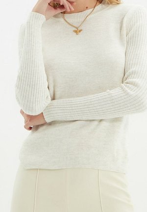 Nainen pukeutunut kermaiseen ribbineulepaitaan, jossa pitkät hihat, ja kultaiseen perhosenmuotoiseen kaulakoruun, poseeraa yhdellä kädellä lähellä kasvojaan.