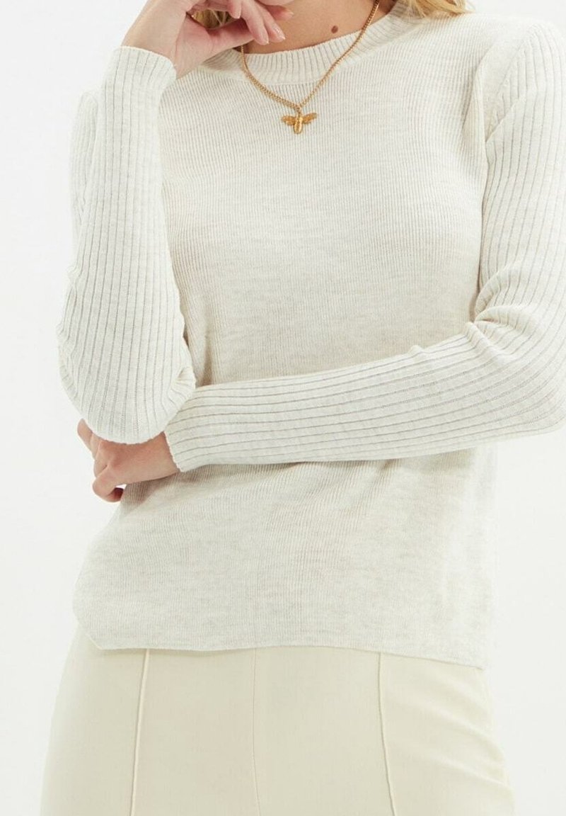 Femme portant un pull côtelé crème à manches longues et un collier papillon doré, posant avec une main près de son visage.