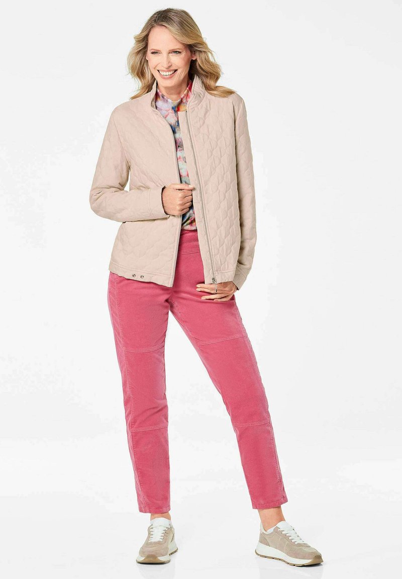 Beige gewatteerde jas met een staande kraag, gedragen over een meerkleurige top en gecombineerd met roze broeken en beige sneakers.