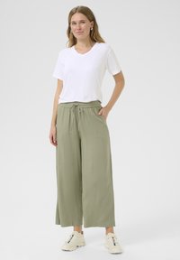 Bela v-neck majica v kombinaciji s širokimi, olivno zelenimi hlacami, ki imajo elastičen pas in sprednje žepke. Športni bež superge.