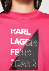 Růžové tričko s krátkým rukávem s rozděleným designem a bílým textem na růžovém a černém pozadí. Materiál vypadá hladce, s kulatým výstřihem.