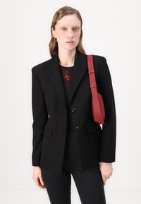 Črn natančno krojen blazer z dvema gumboma, zareznimi ovratniki in žepi, v kombinaciji s topom okroglega ovratnika z cvetličnim vzorcem in rdečo torbico na čez ramo.