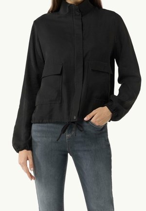 Frau trägt eine schwarze langärmelige Jacke mit vorderen Klapptaschen und Kordelzug in der Taille, kombiniert mit blauen Jeans.