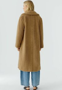 Vrouw in een lange bruine teddyjas, brede blauwe jeans en zwarte open hakken, staand en met haar rug naar de camera gekeerd tegen een effen achtergrond.
