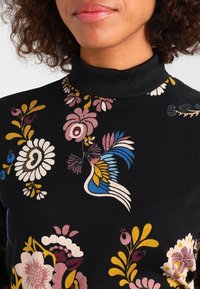 Svart polotröja med flerfärgad broderi av blommor och fåglar i rosa, gult, blått och vitt. Mjukt tyg med en slät yta.