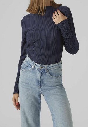 Vrouw draagt een marineblauw geribd shirt met lange mouwen, gestopt in een lichtblauwe high waist jeans, tegen een effen achtergrond.