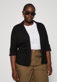 Blazer nero con maniche arrotolate, abbinato a una camicia bianca e pantaloni cargo verde oliva. Il modello indossa occhiali da sole scuri e orecchini a cerchio.