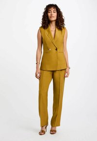 Chaleco sastre de color mostaza, con blazers sin mangas de doble botonadura y pantalones de pierna ancha, textura suave, diseño minimalista.