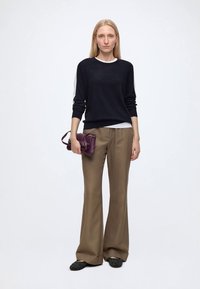 Femme debout aux longs cheveux blonds portant un pull noir, un pantalon beige à jambes larges, des chaussures noires, et tenant un petit sac à main violet.