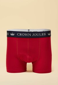 Red cotton boxer shorts with a navy elastic waistband featuring the text "CROWN JOULES" and a crown logo. Smooth texture and fitted design.