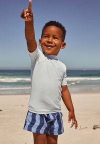 Maglia da bagno a maniche corte azzurro chiaro e pantaloncini da nuoto a righe blu navy. Bambino indica, con mano sabbiosa, davanti a uno sfondo di spiaggia.