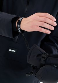 Svart skidjacka med en SLOPE®-logga, som visar en hand som håller ett svart rem med SLOPE®-branding, fäst vid matchande handskar.
