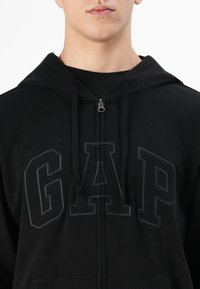 Close-up of a person wearing a black zip-up hoodie with a hood and "GAP" embroidered in large black letters across the chest.