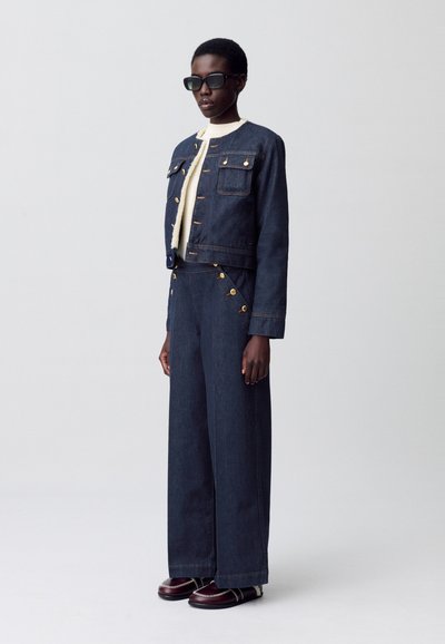 Giacca di jeans con bottoni d'oro e due tasche sul petto, abbinata a pantaloni in denim a gamba larga. Il modello indossa occhiali da sole scuri e scarpe bordeaux.