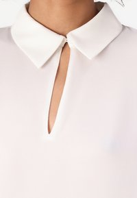 Blusa blanca con cuello, que presenta un escote en forma de cerradura. Tela suave con una textura lisa, diseño minimalista y un brillo sutil.