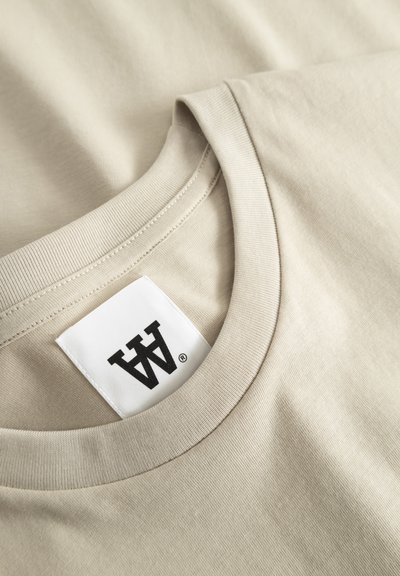 T-shirt en coton beige avec un col rond côtelé et une étiquette carrée avec un logo noir brodé à l'arrière. Texture douce et lisse.