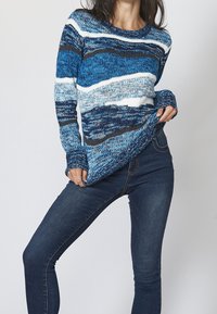 Blå stickad tröja med vågiga mönster i ljus och mörk blå, med långa ärmar och rund halsringning, tillsammans med mörka denimjeans.