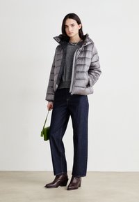Doudoune grise avec capuche, fermeture éclair à l'avant et design matelassé horizontal. Associée à un pull gris, un jean foncé et des bottines brunes.