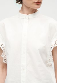 Femme portant une chemise blanche à boutons avec un col mandarin et des détails en dentelle sur les manches courtes, légèrement tournée vers la droite.