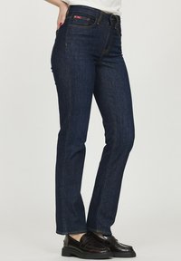 Jeans en denim bleu marine avec une coupe droite, des coutures jaunes et cinq poches, associés à des mocassins noirs.