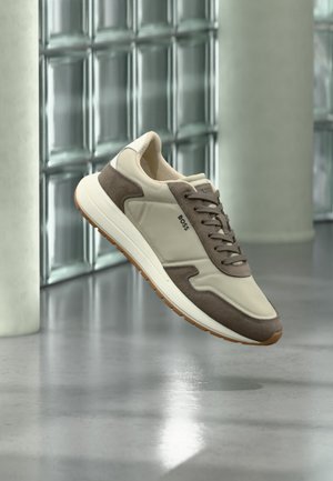 Beiger und brauner BOSS Sneaker mit Wildledereinsätzen, Schnürsenkeln und Gummisohle, schwebend über poliertem Betonboden nahe einer Glassteinwand.