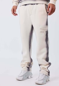 Persona con pantaloni della tuta bianchi con un logo ricamato sulla coscia e scarpe da ginnastica bianche, in piedi su uno sfondo chiaro e uniforme.