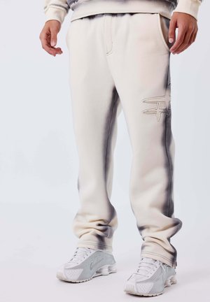 Persoon in witte joggingsbroek met een gestikt logo op de dij en witte sport sneakers, staand op een effen lichte achtergrond.