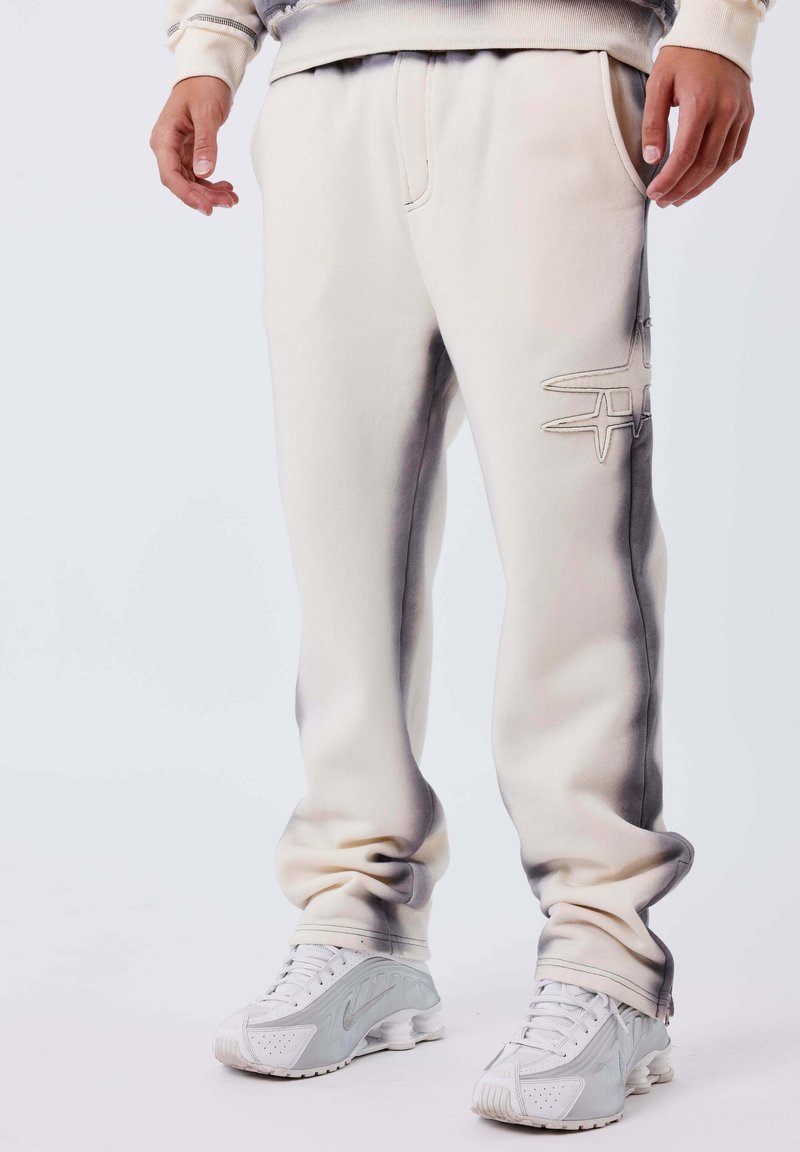 Persona con pantaloni della tuta bianchi con un logo ricamato sulla coscia e scarpe da ginnastica bianche, in piedi su uno sfondo chiaro e uniforme.