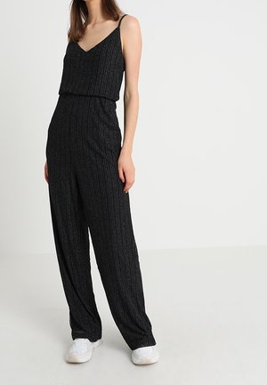 Schwarzer Jumpsuit mit dünnen Trägern, gerippter Textur und lockeren, weiten Beinen. Kombiniert mit weißen Sneakers für den Kontrast.