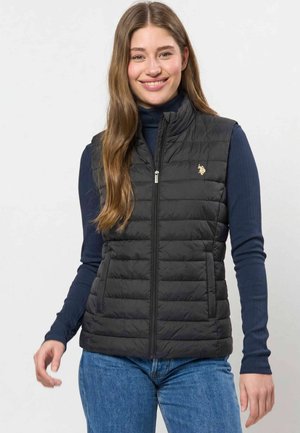 Jeune femme aux longs cheveux bruns souriante, portant une veste noire matelassée sur un t-shirt côtelé bleu marine à manches longues et un jean bleu.
