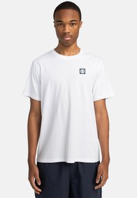 Camiseta de algodón blanca con mangas cortas, cuello redondo y un pequeño parche de logo azul en el lado izquierdo del pecho. Corte relajado.