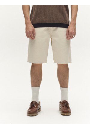 Mees, kandes beežid põlveküpsed lühikesed püksid, valged randmesokid, pruunid nahast kingad ja tume kootud särk.