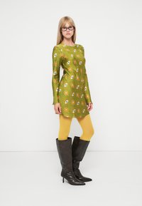Vestido verde de mangas compridas com padrão floral, collants amarelos e botins castanhos até ao joelho com salto. Tecido suave, design ajustado e cores vibrantes.