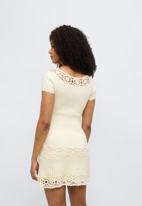 Πλεκτό φόρεμα σε ανοιχτό κρεμ χρώμα, με κοντά μανίκια, σκουλικωτή λαιμόκοψη κροσέ και δαντέλα στο τελείωμα με υφή.