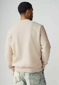 Beige huppari, jossa on raidoitettu kaula-aukko ja hihansuut, sileä pinta ja rento leikkaus, yhdistettynä vaaleansinisiin kulutettuihin farkkuihin.