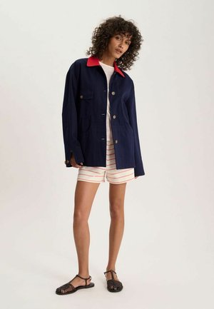 Femme aux cheveux bouclés portant une veste bleu marine boutonnée avec col rouge, chemise blanche, short rayé et sandales noires tressées, debout.