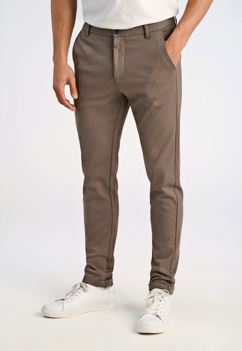 Pantaloni slim-fit in tessuto taupe, caratterizzati da una superficie liscia, tasche frontali e dettagli di cucitura a contrasto. Abbinati a sneakers bianche.