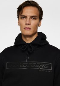 Musta huppari, jossa on naru hupussa ja teksturoitu, sävyyn sopiva logo rinnassa. Sujuva kangas rentoa istuvuutta varten.