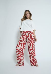 Weit geschnittenes weißes Baumwoll-T-Shirt, kombiniert mit weit geschnittenen Hosen mit einem roten Wellenmuster auf cremefarbenem Hintergrund. Schlichte Riemchensandalen.